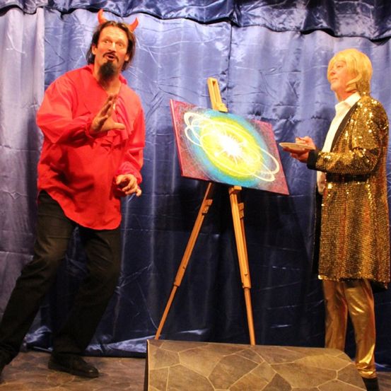 Zwei Darsteller auf der Bühne. Einer trägt ein rotes Shirt und Teufelshörner und gestikuliert lebhaft. Der andere, in Goldpailletten, hält einen Teller in der Nähe eines Staffeleis mit einem abstrakten Gemälde.