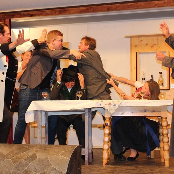 Eine Gruppe von Menschen führt ein Sketch auf einer Bühne auf, wobei zwei Männer versuchen, einem sitzenden Mann den Hut abzunehmen. Eine weitere Person gestikuliert, während andere Weinglas halten.