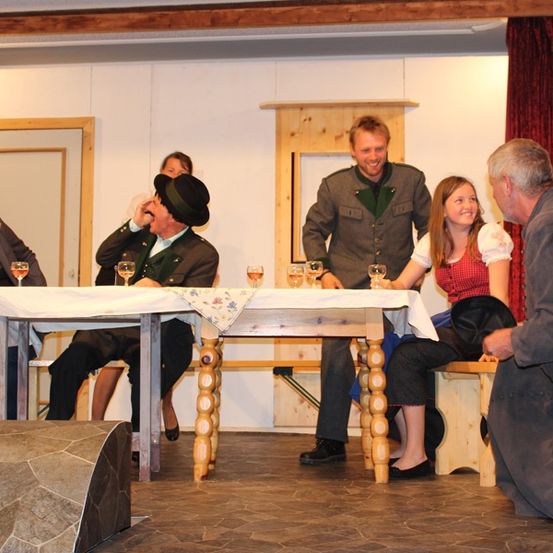 Sechs Personen sitzen um einen Tisch herum, einige trinken Wein. Ein Mann steht hinter dem Tisch und ein anderer Mann kniet in der Nähe. Die Umgebung scheint eine inszenierte Szene zu sein.