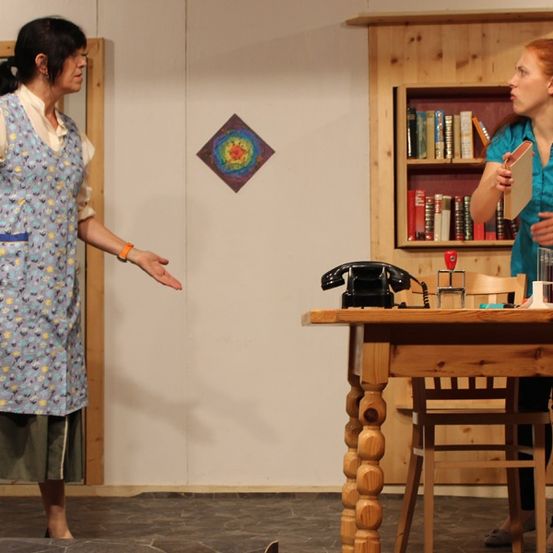 Zwei Frauen stehen auf einer Bühne. Eine zeigt mit der Hand, während die andere ein Buch hält. Hinter ihnen steht ein Tisch mit Büchern, einem Telefon und einem Glaskrug.