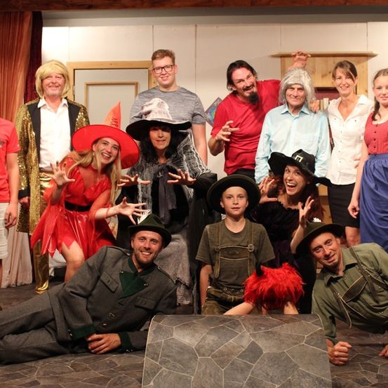 Eine Gruppe von Menschen in Kostümen, die für ein Foto posieren, mit einer Person auf dem Boden. Einige tragen Hüte, Brillen und Stiefel. Der Hintergrund besteht aus einer Holzwand und einer Tür.