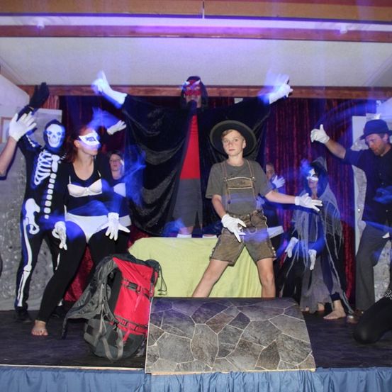 Eine Gruppe von Menschen auf der Bühne, einige in Kostümen, einer in einem Skelettkostüm, ein anderer in einem schwarzen Umhang, andere in weißen Handschuhen, mit einem roten Rucksack auf dem Boden.