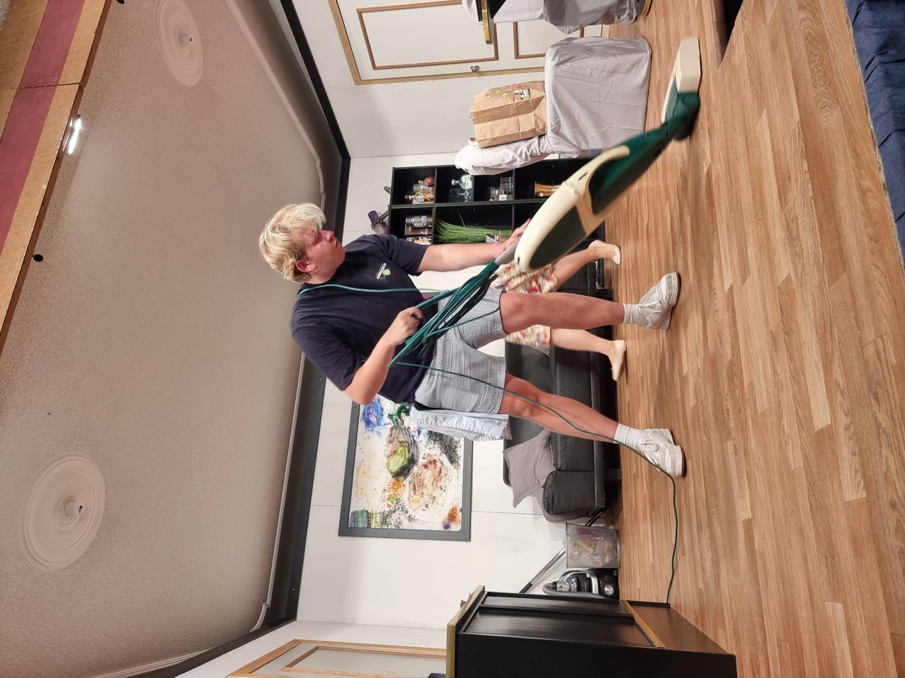 Ein Mann in einem blauen T-Shirt und Shorts benutzt einen Mop, um den Boden zu reinigen, während er ein Kabel in der rechten Hand hält.