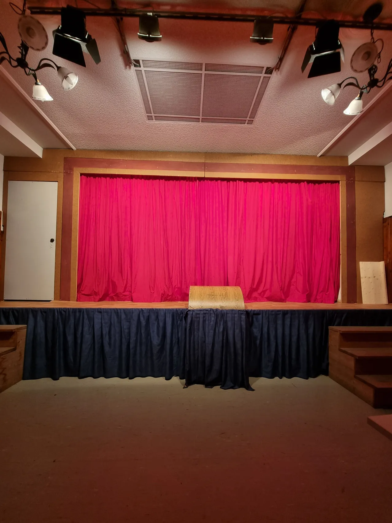 Eine leere Theaterbühne mit rotem Vorhang und blauen Vorhängen, einem hölzernen Stuhl auf der Bühne und hölzernen Stufen an den Seiten.