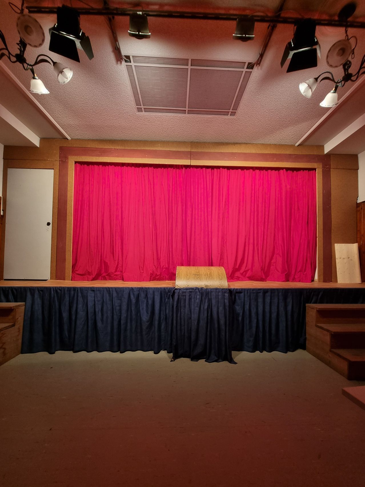 Eine leere Theaterbühne mit rotem Vorhang und blauen Vorhängen, einem hölzernen Stuhl auf der Bühne und hölzernen Stufen an den Seiten.