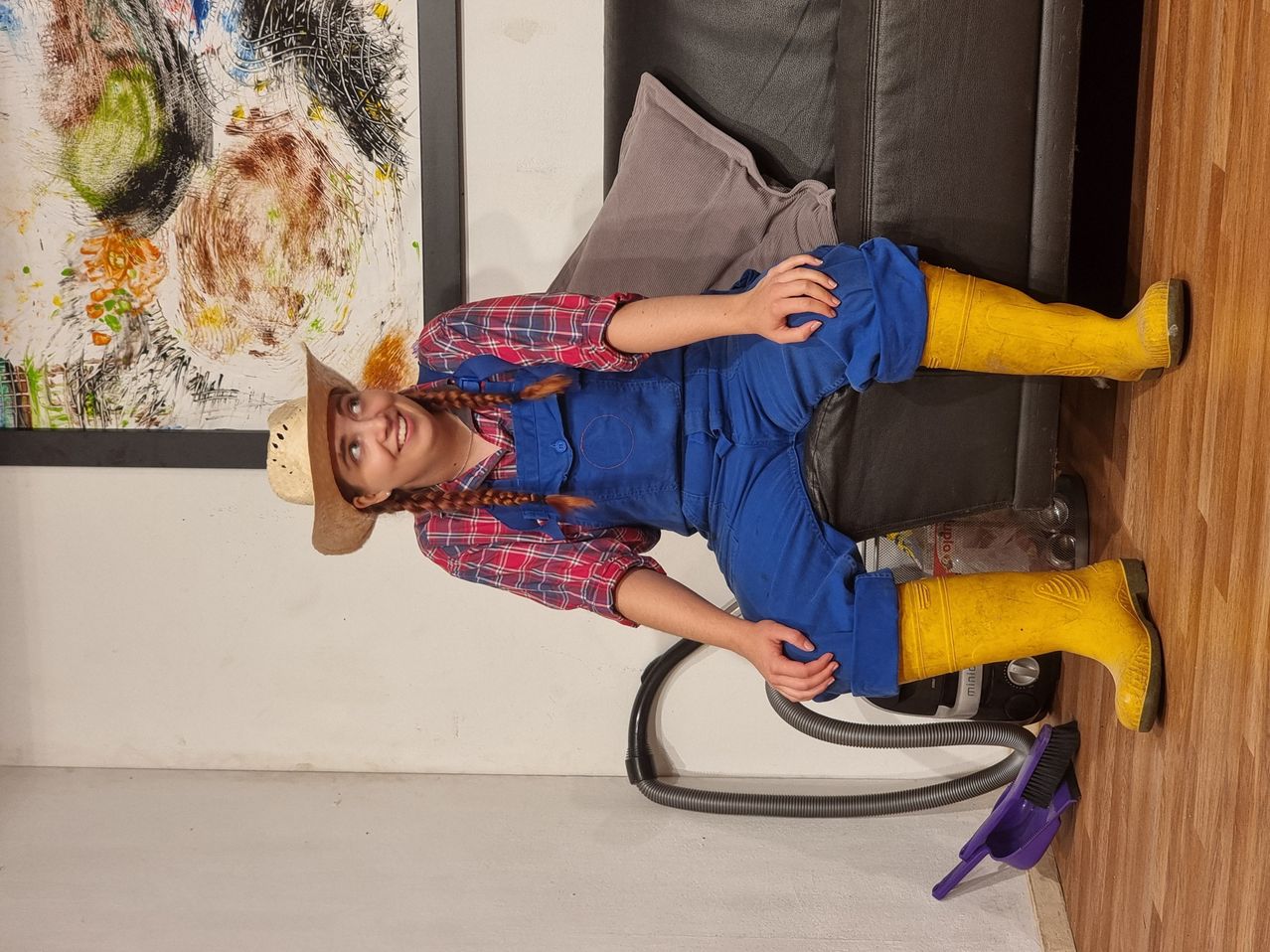 Eine Frau liegt mit blauer Latzhose und gelben Stiefeln auf einem schwarzen Sofa und lächelt, mit einem Pinsel und Staubsauger in der Nähe.