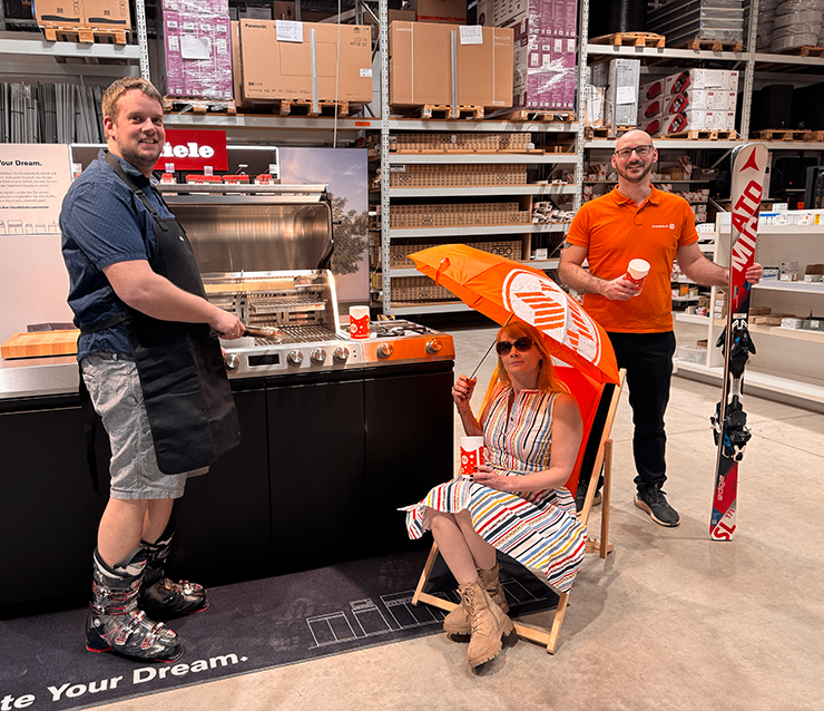 Three people in a warehouse. A man with an apron stands near a grill, a woman with an umbrella sits in a chair, and a man in orange holds a ski.