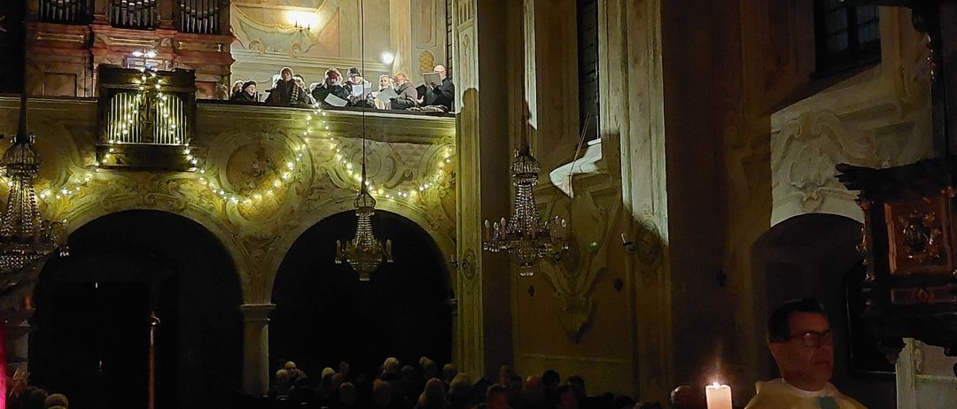 Ein Priester steht vor einem Altar in einer Kirche, umgeben von Kerzen. Die Gemeinde sitzt auf Kirchenbänken mit Lichtern und Dekorationen. Menschen befinden sich auf der Galerie oben.