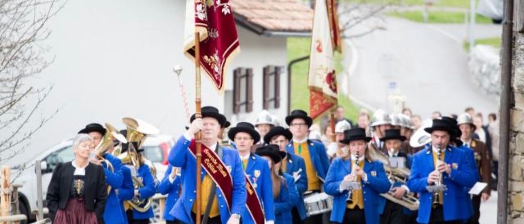 Eine Musikkapelle in blauen Uniformen und Hüten, die Musikinstrumente halten, geht vor einem Haus mit gefliestem Dach die Straße entlang.