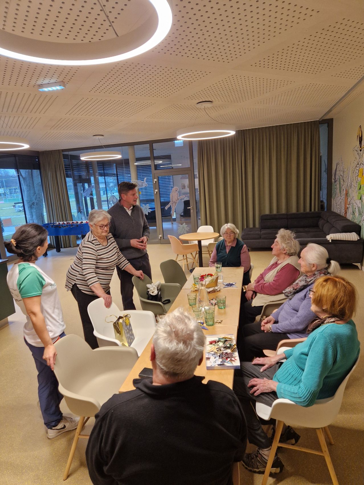 Eine Gruppe älterer Frauen und Männer versammelt sich um einen Tisch in einem Raum. Eine ältere Frau greift nach einer Geschenktüte auf dem Tisch. Der Raum hat weiße Stühle, einen runden Tisch mit Gläsern und einer Schüssel und eine Couch in der Nähe der Glastür.