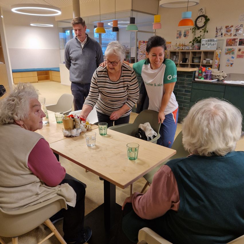 Mehrere ältere Menschen sitzen um einen Tisch in einem Raum. Eine Frau mit Brille steht neben einer älteren Frau und lächelt. Eine andere Frau schaut zu.