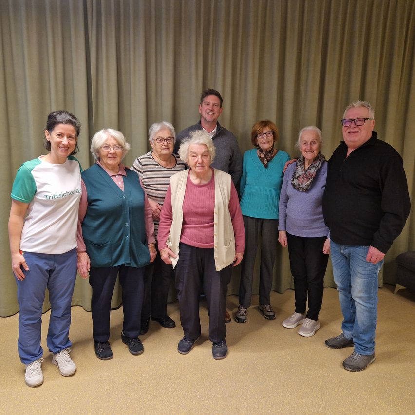 Eine Gruppe älterer Menschen und zwei Männer stehen vor einem Vorhang und posieren für ein Foto. Die Menschen tragen Freizeitkleidung, Brillen und einige haben Schals um. Der Boden ist mit Teppich ausgelegt.