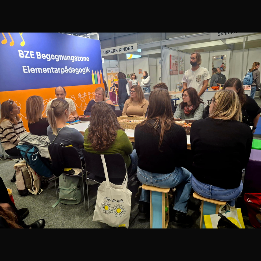 Eine Gruppe von Menschen sitzt an einem Tisch auf einer Messe, in einer Diskussion vertieft. Ein Banner im Hintergrund lautet 'BZE Begegnungszone Elementarpädagogik'. Im Hintergrund befinden sich verschiedene Menschen, einige stehend, andere sitzend.