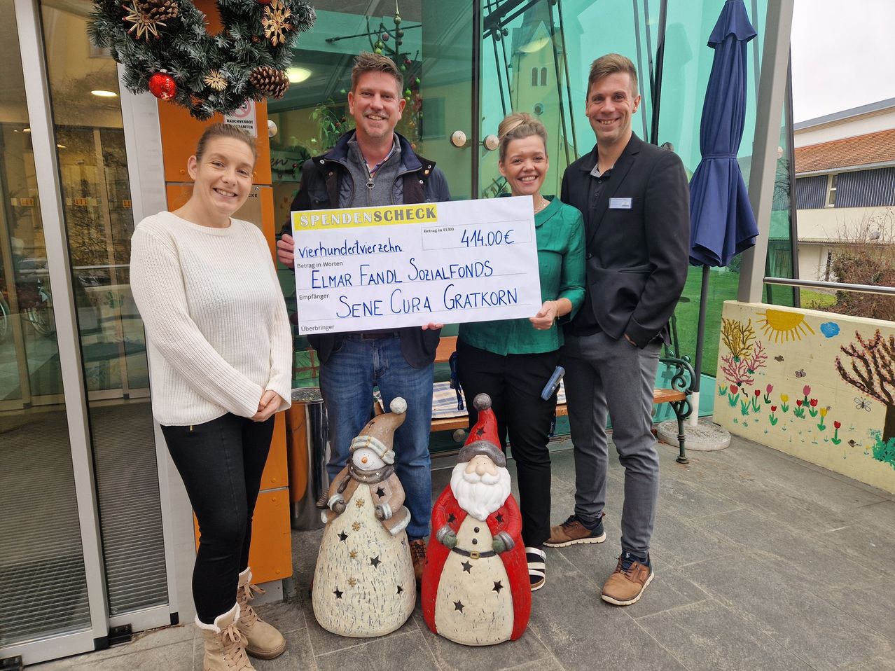 Vier Personen stehen vor einem Gebäude und halten einen Spendenscheck. Der Scheck beträgt 444 Euro. Zwei Männer und zwei Frauen lächeln. Hinter ihnen stehen zwei Schneemänner. Rechts befindet sich ein blauer Regenschirm.