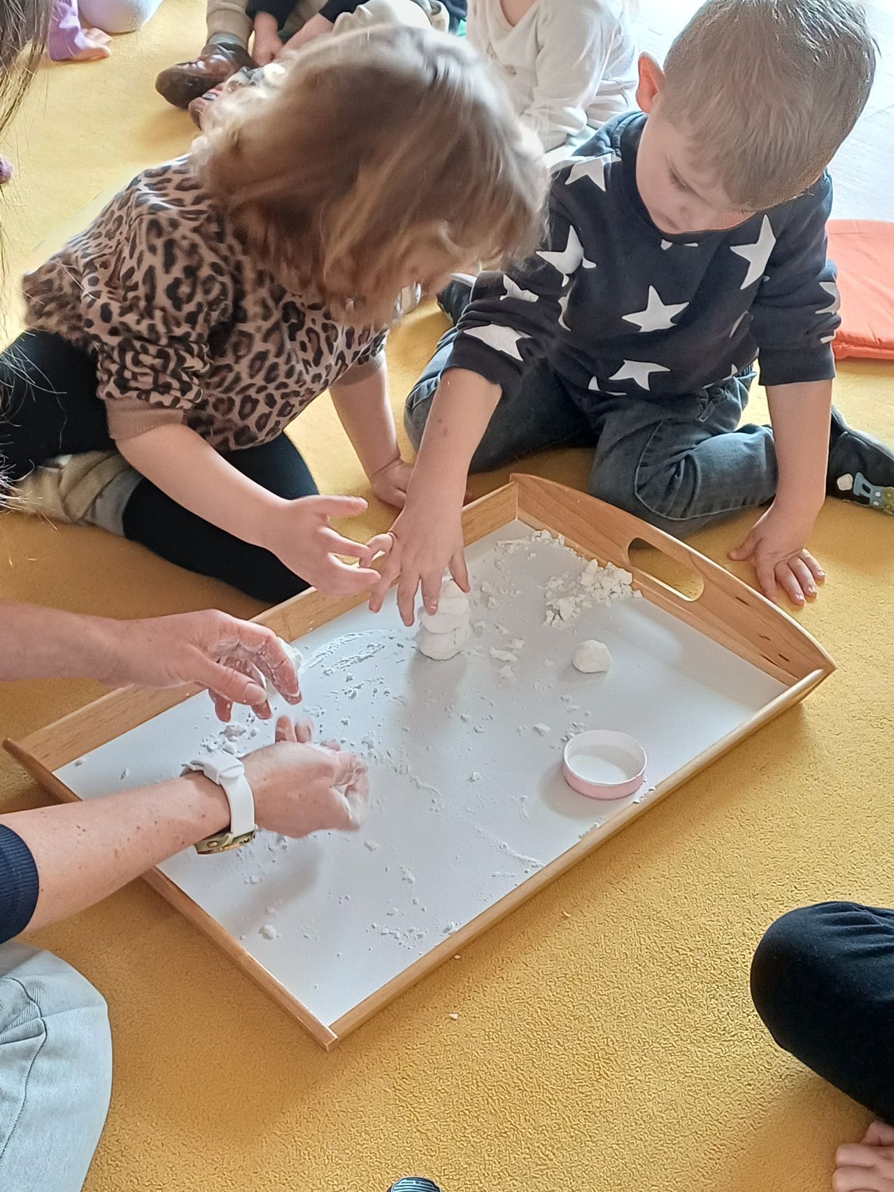 Zwei Kinder und ein Erwachsener spielen auf einem Holztablett mit weißer Pulver. Das Kind auf der linken Seite trägt ein Leopardenmusterhemd und schwarze Hose. Das Kind auf der rechten Seite trägt ein schwarzes Shirt mit weißen Sternen und blaue Jeans.