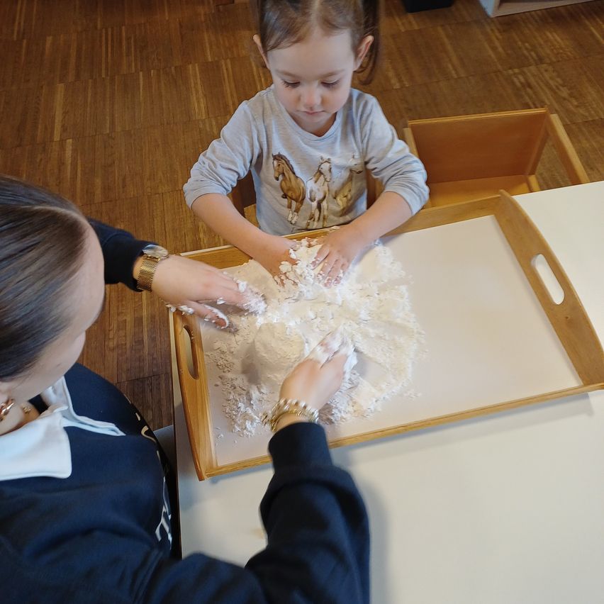 Ein junges Mädchen und eine erwachsene Frau spielen mit weißem Pulver auf einem Tablett in einem Raum.