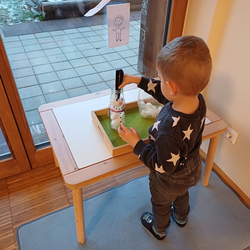 Ein junger Junge steht an einem Tisch und spielt mit einem Schneemann in einem Glas. Er hält einen Stock und ein Glas voller Wattebälle. Er scheint einen Schneemann zu basteln.