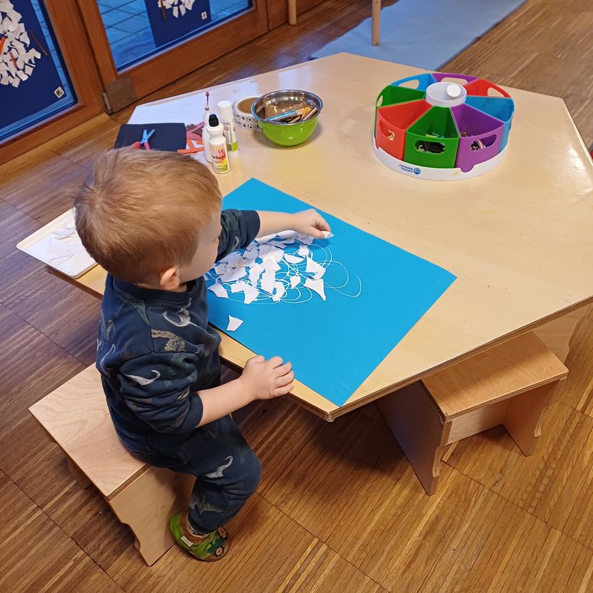 Ein junger Junge sitzt an einem Tisch und arbeitet an einer blauen Papierbastelei mit verschiedenen Ausschnitten. Ein bunter Spielzeugorganizer steht auf dem Tisch, und es gibt mehrere Bastelutensilien in der Nähe.