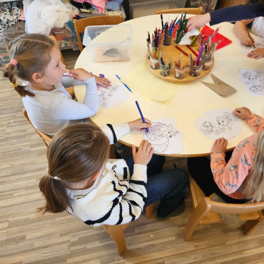 Several young girls are seated around a table, engaged in coloring activities. A variety of colored pencils are placed on the table, and the floor is wooden.