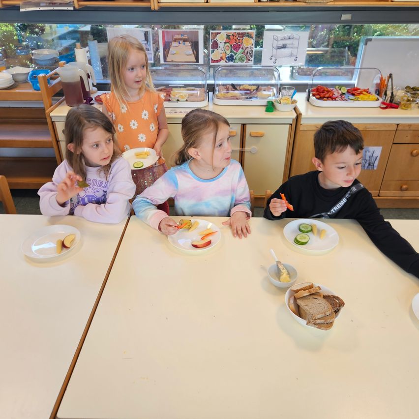 Four children sit at a table with plates of food, one with an apple slice. A girl stands behind them, looking at another girl. Trays with food are on the counter.