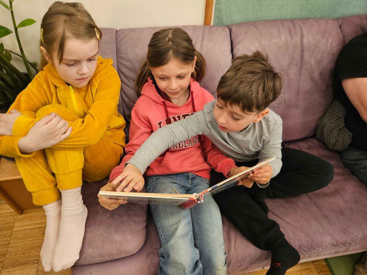 Three children sit on a purple couch, one reading a book while the others watch. The girl in yellow is looking away, and the other girl smiles. The boy points at the book.