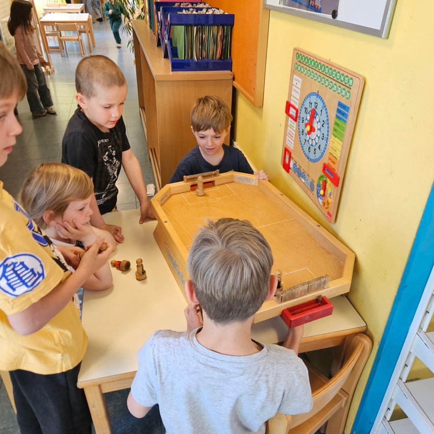 Several young boys are playing a board game in a classroom. A clock and alphabet chart is on the wall behind them.