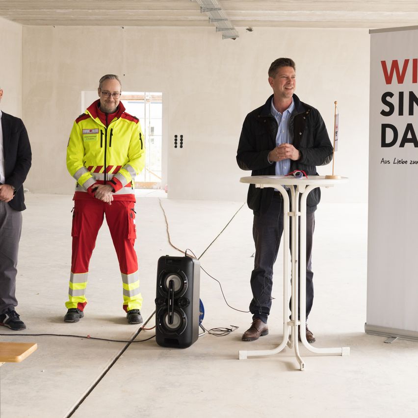 Drei Männer stehen in einem unfertigen Raum. Ein Mann in einer reflektierenden Weste steht neben einem Lautsprecher, ein anderer Mann spricht an einem Podium.