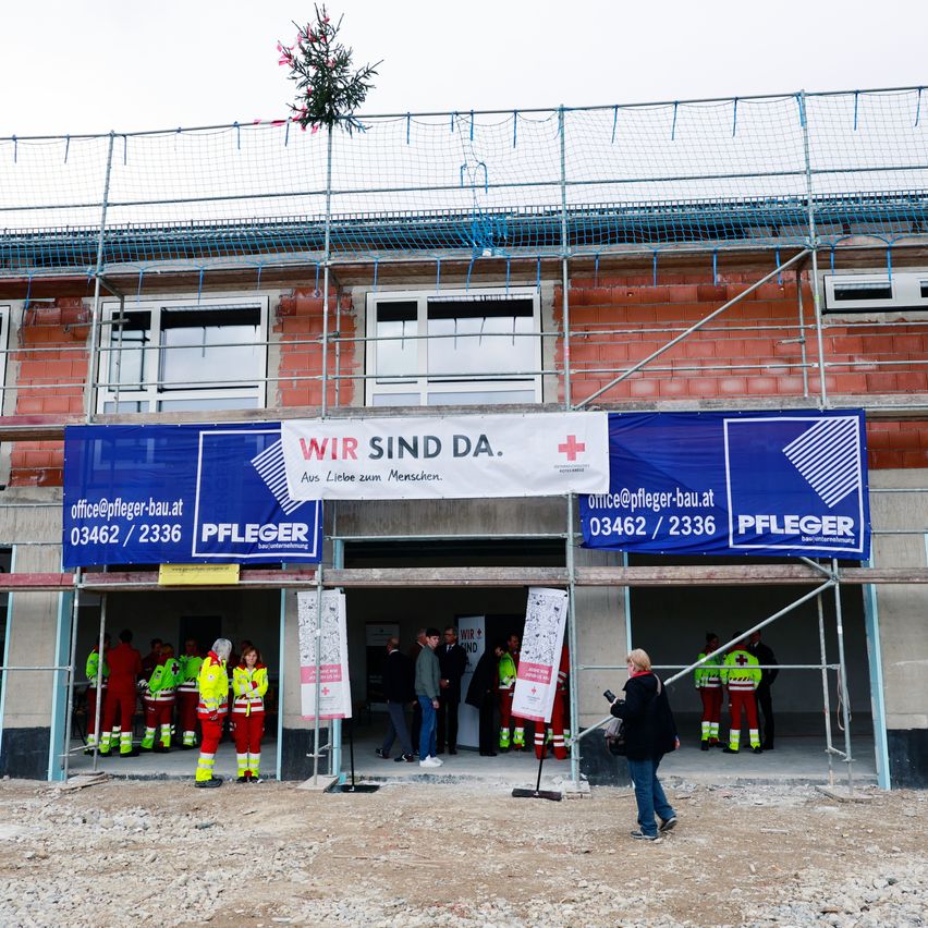 Ein im Bau befindliches Gebäude mit blauen Werbebannern von Pfleger Bau. Mehrere Personen in roten und gelben Uniformen stehen vor dem Eingang.