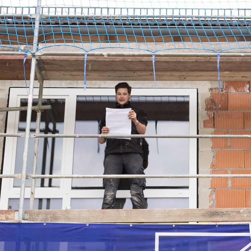 Ein Mann steht auf einem Gerüst, hält ein Stück Papier, möglicherweise einen Bauplan, in der Hand und sieht auf ein Fenster. Das Gerüst ist mit einem blauen Netz bedeckt.
