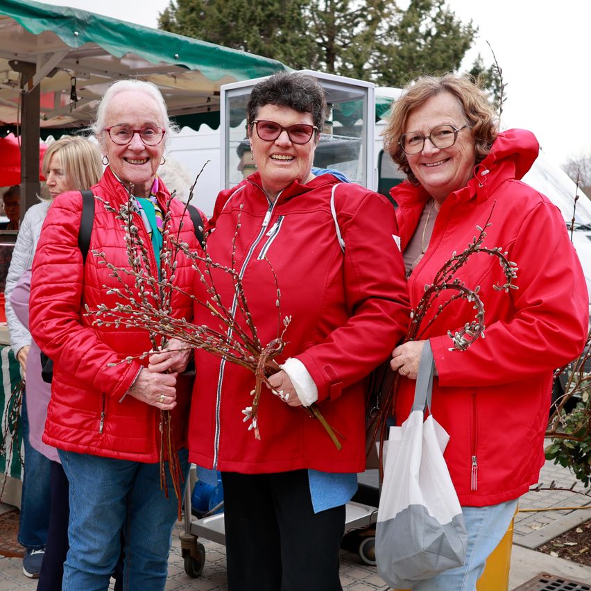 Drei Frauen in roten Jacken lächeln für ein Foto und halten Stöcke mit Blättern auf einem Markt. Hinter ihnen sind ein grüner Vordach und ein weißer Van zu sehen.