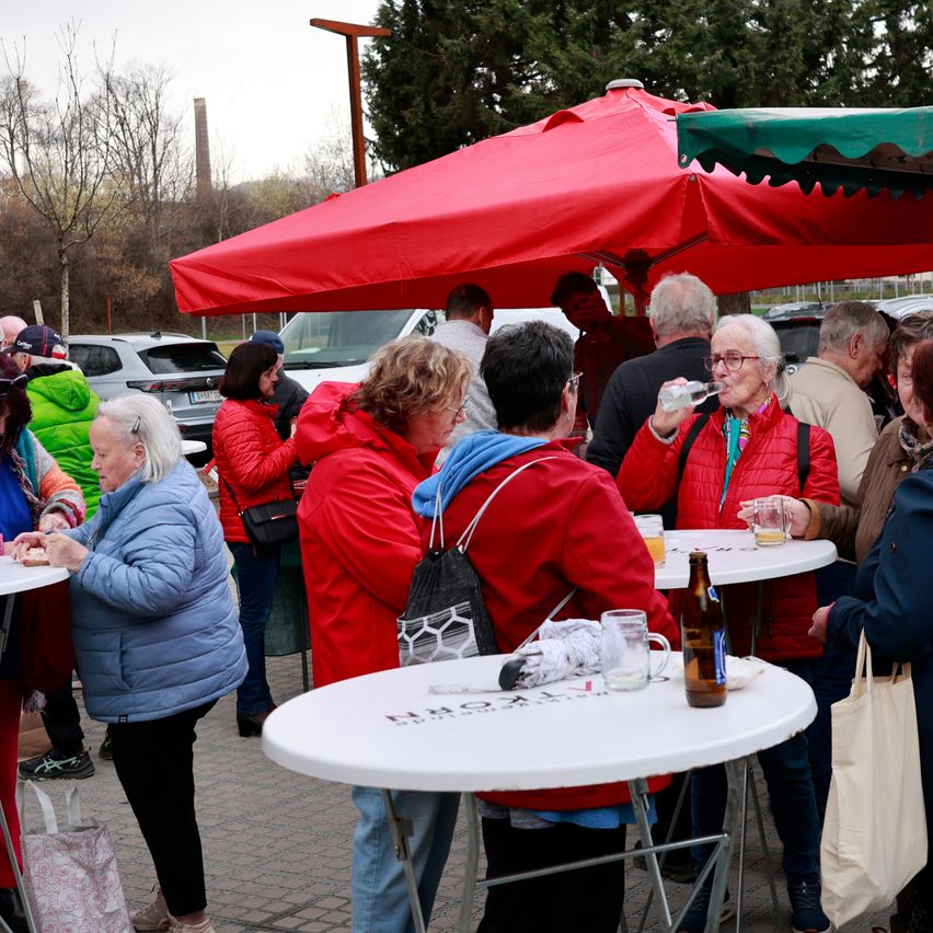 Eine Gruppe von Menschen sitzt um Tische unter einem roten Zelt in einem Park. Einige trinken und andere essen. Eine Frau trägt eine Brille und trinkt aus einer Flasche. Hinter ihnen stehen Bäume und geparkte Autos.