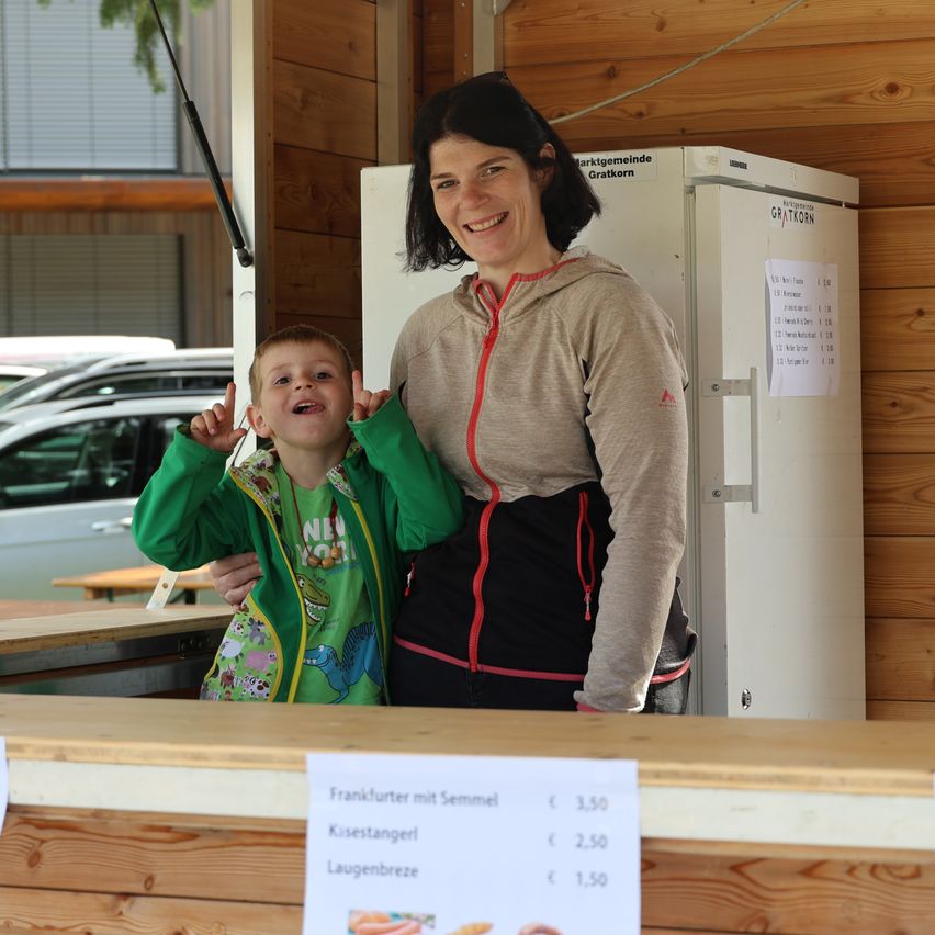 Eine Frau und ein kleiner Junge stehen lächelnd vor einer Theke und posieren für ein Foto. Die Theke hat eine Holzoberfläche und zeigt ein Menü mit Preisen.