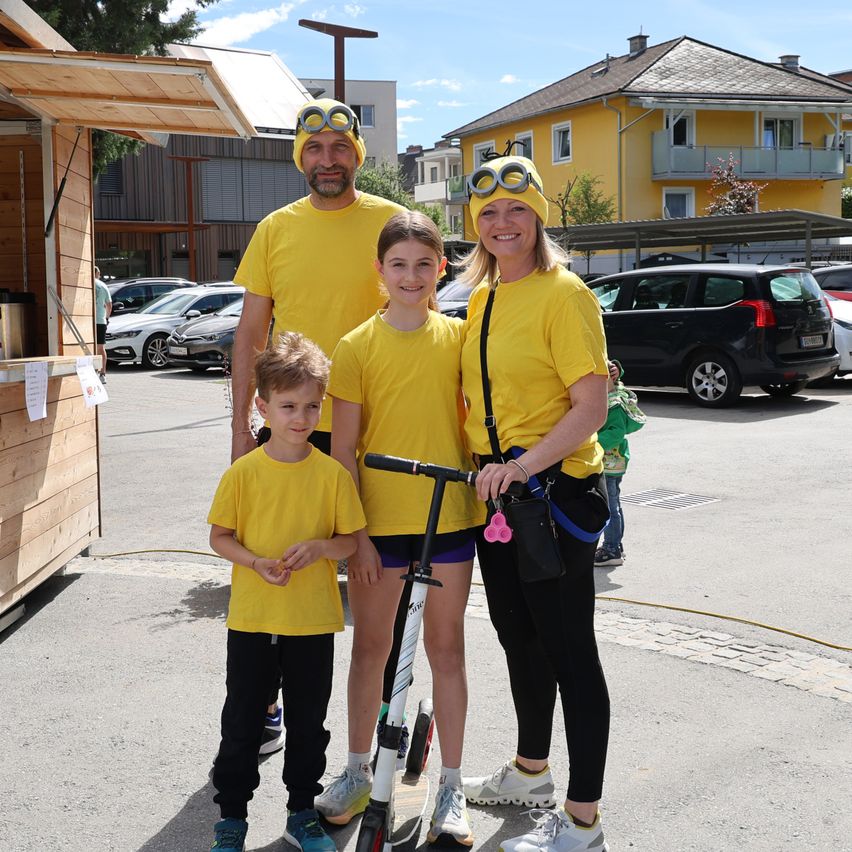Eine Familie in passenden gelben Outfits steht in einem Parkplatz mit einem Roller. Hinter ihnen sind ein Gebäude, Autos und ein Baum sichtbar.