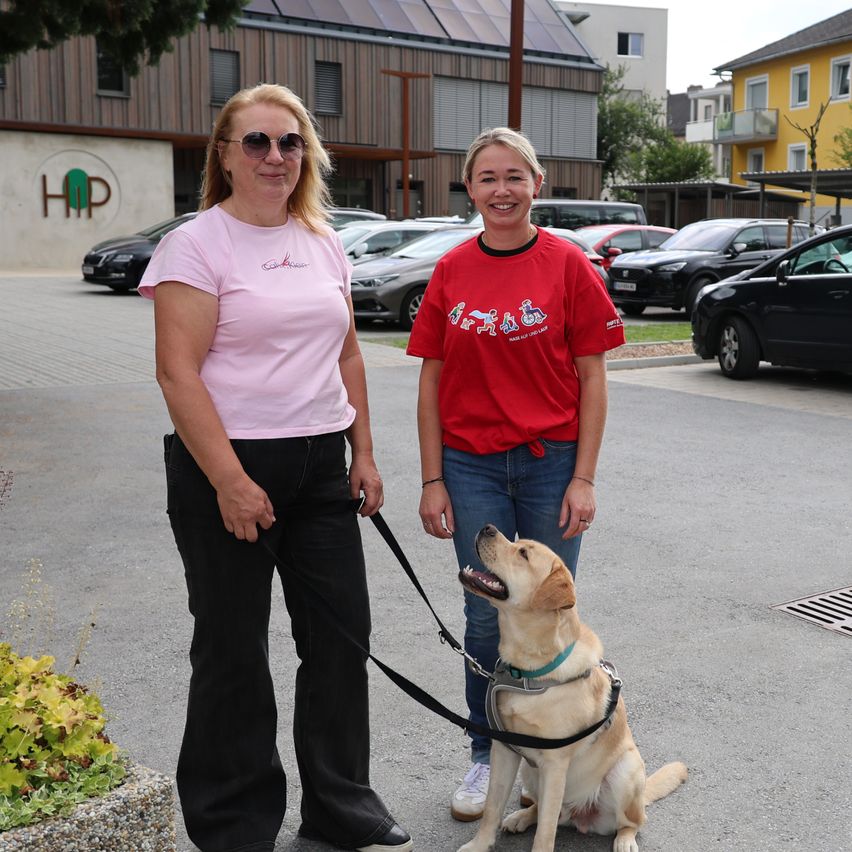 Zwei Frauen stehen mit einem Hund auf einem Parkplatz, mit Gebäuden und Autos im Hintergrund.