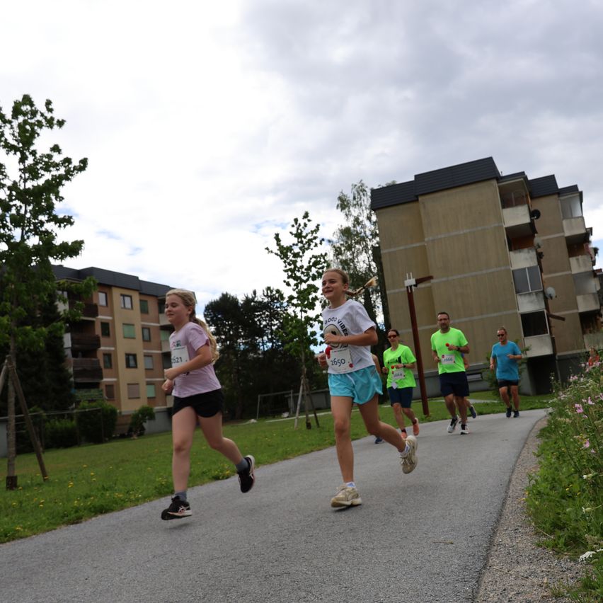 Eine Gruppe von Menschen läuft auf einer asphaltierten Straße einen Marathon. Sie haben Nummern auf ihren Trikots. Gebäude befinden sich an der Seite.