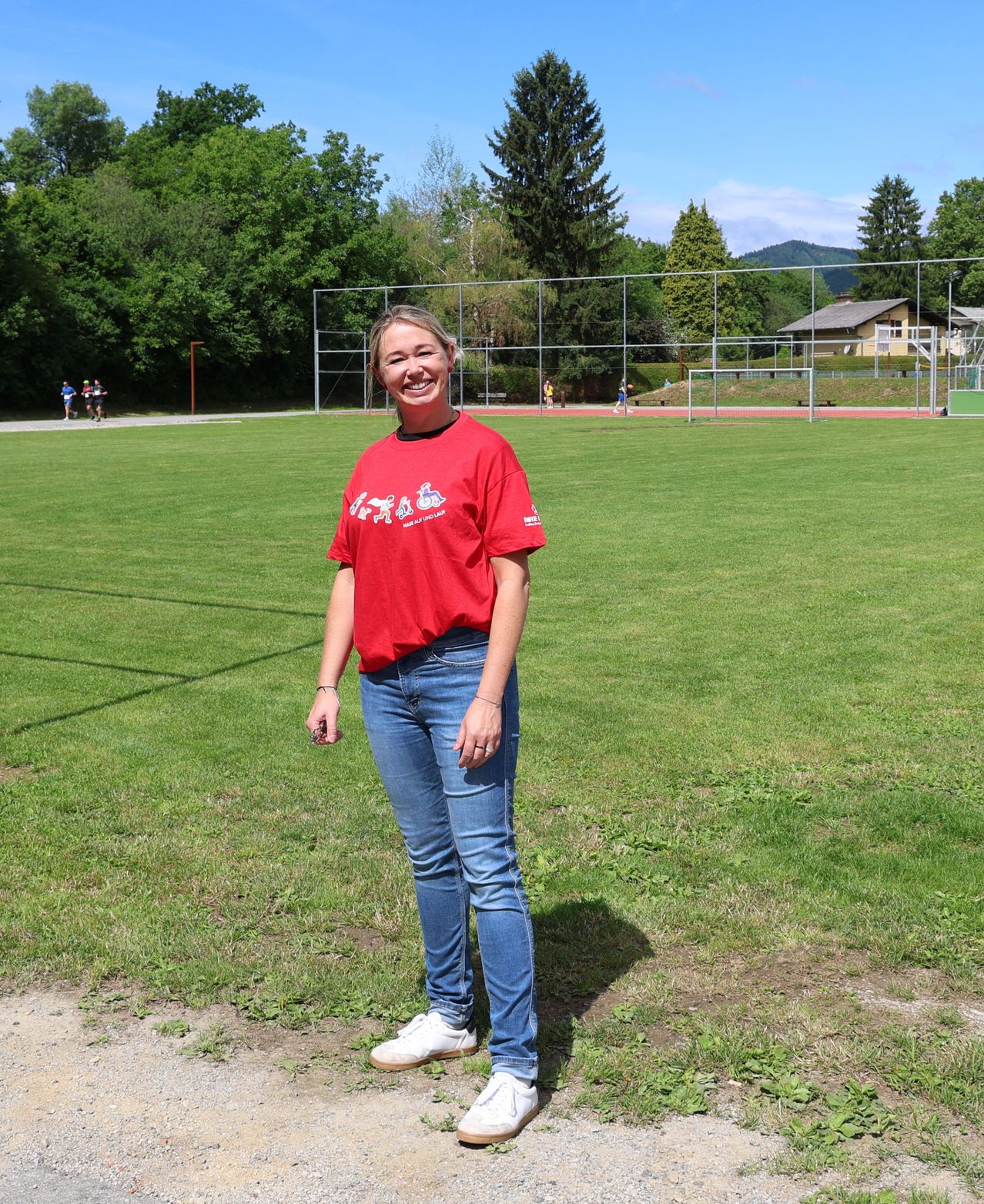 Eine Frau in einem roten T-Shirt steht auf einem Rasenfeld mit einer Sportanlage im Hintergrund.