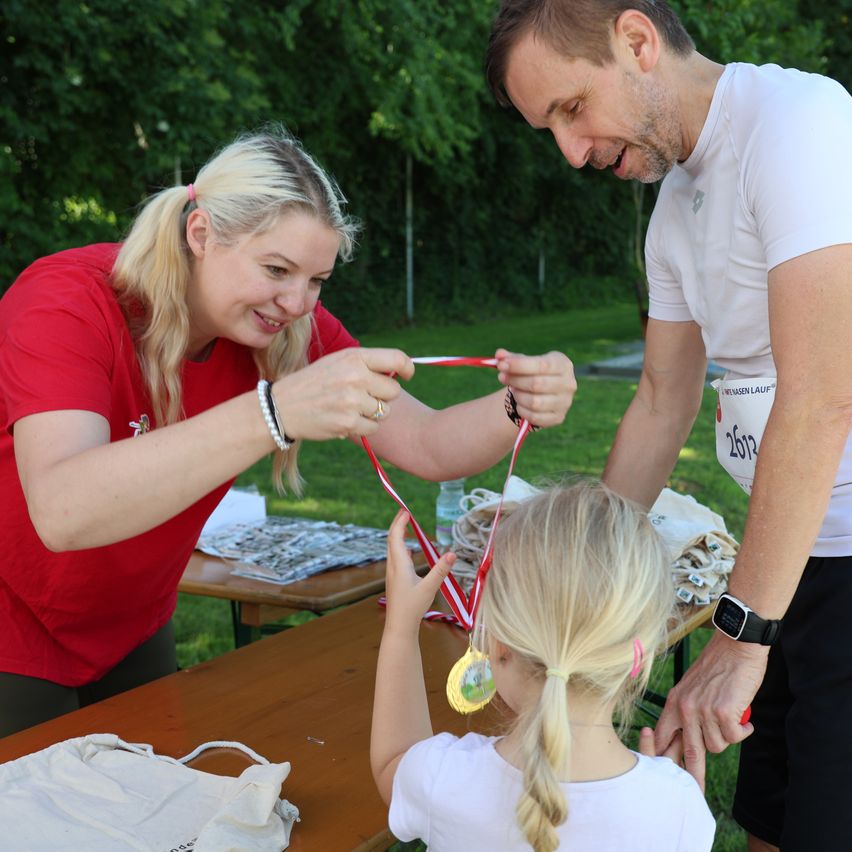 Eine Frau in einem roten T-Shirt legt eine Medaille um den Hals eines jungen Mädchens, während ein Mann in der Nähe lächelt. Sie befinden sich in einer Parkumgebung mit Bäumen und einem Tisch mit Gegenständen im Hintergrund.