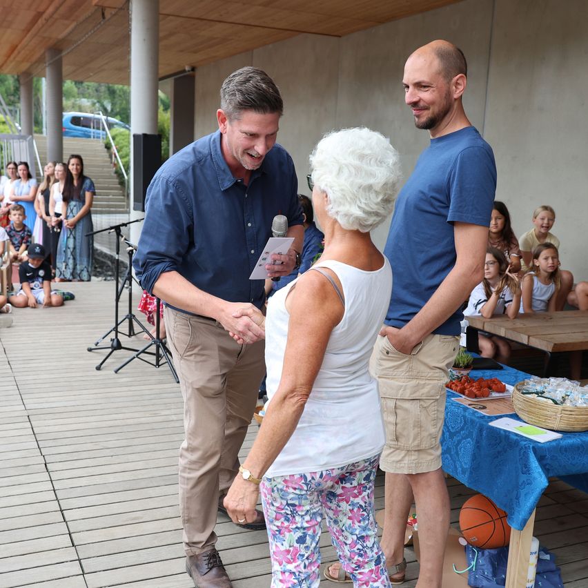 Two men and an elderly woman stand on a deck, shaking hands. A microphone and paper are in the man's hand. A table with food and a basketball is behind them, and a crowd is watching.