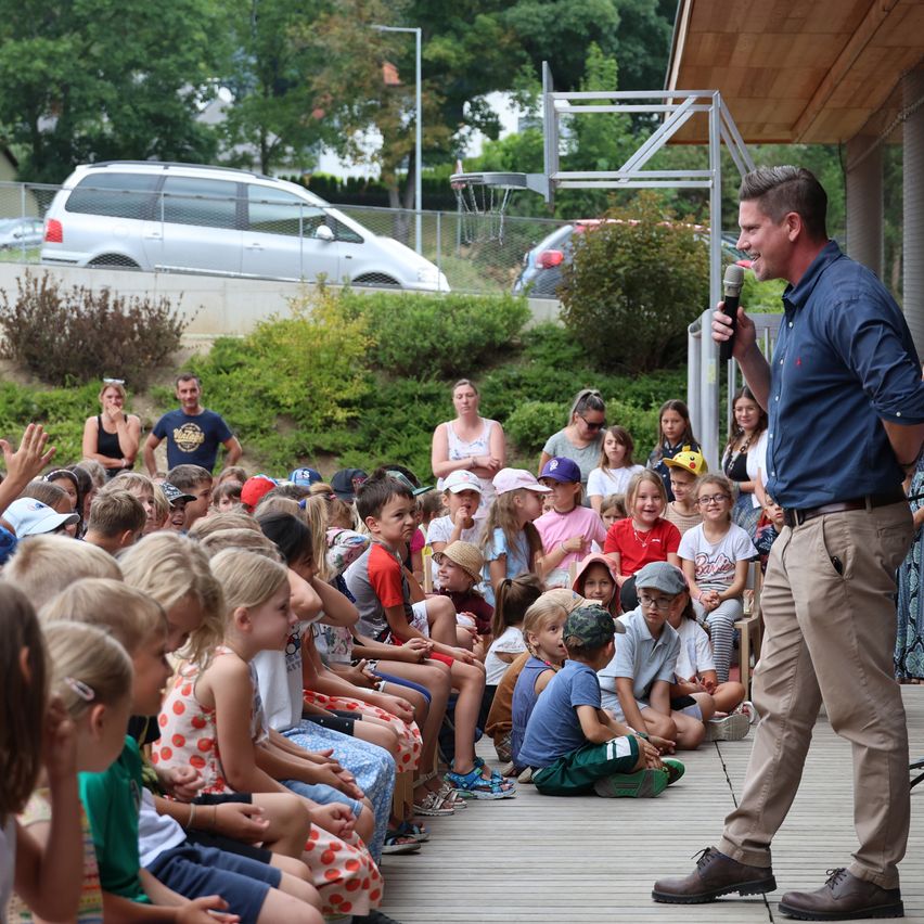 A man stands on a stage, holding a microphone, and addresses a group of children sitting on the ground. The children are attentively listening to him. Behind the man, there are several adults standing and sitting on the grass. In the background, there are bushes, a fence, and a parked car.