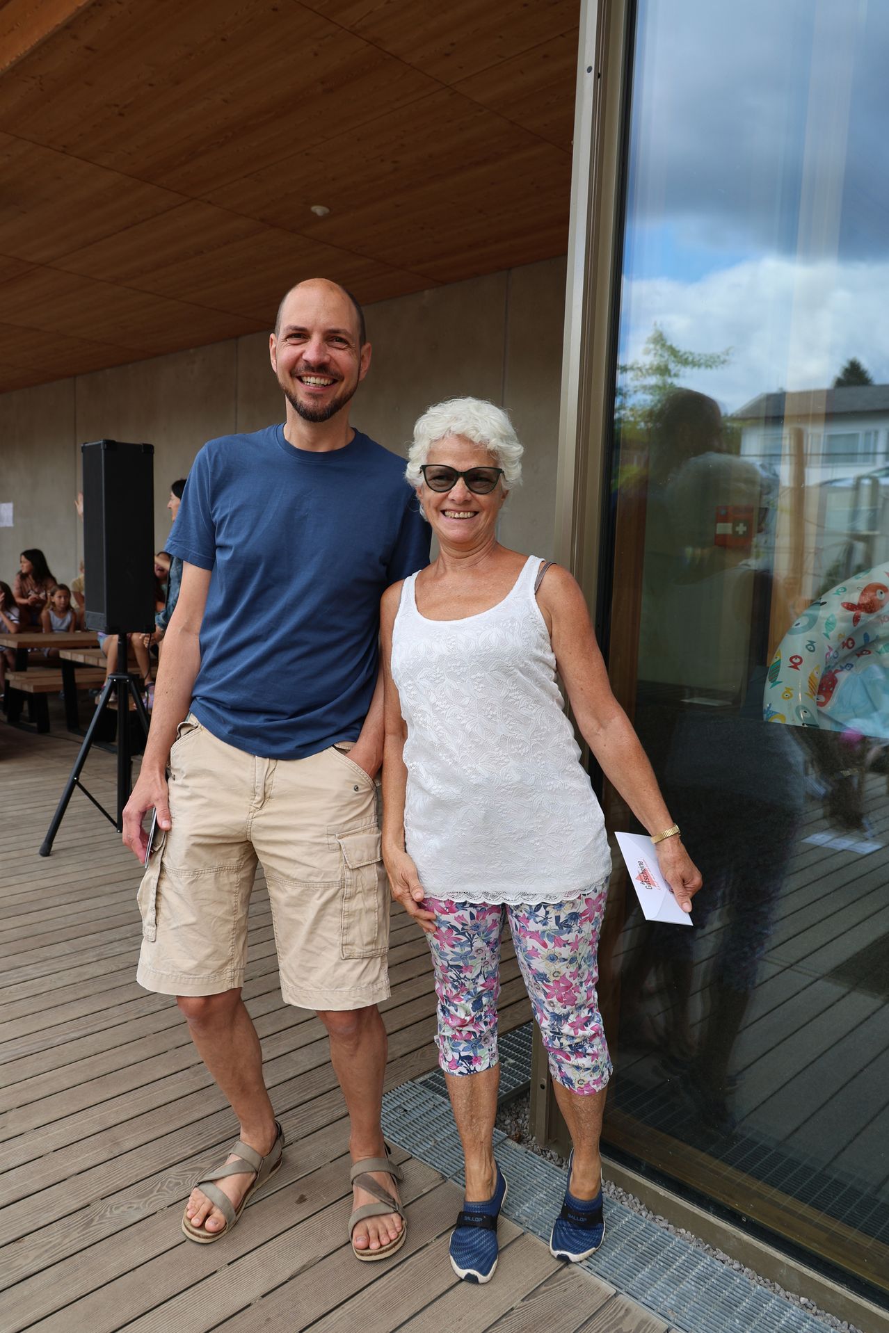 A man and an older woman stand together on a deck, smiling and posing for a photo. The man wears a blue shirt and khaki shorts, while the woman dons a white tank top and floral pants. Behind them, a glass wall reflects a blurred scene of people and a speaker.