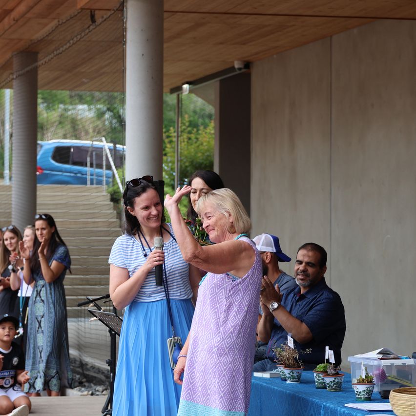 A woman is being honored at an outdoor event. She is standing with a woman holding a microphone. Behind them, a group of people are clapping. In the background, there is a staircase and a blue van.