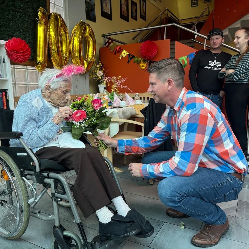 A man in a plaid shirt kneels to give a bouquet of flowers to a woman in a wheelchair, celebrating her 100th birthday with balloons and a festive backdrop.