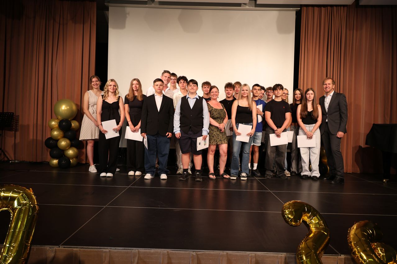 A group of young people stand on stage holding certificates, smiling for a photo. Behind them is a white screen and balloons.