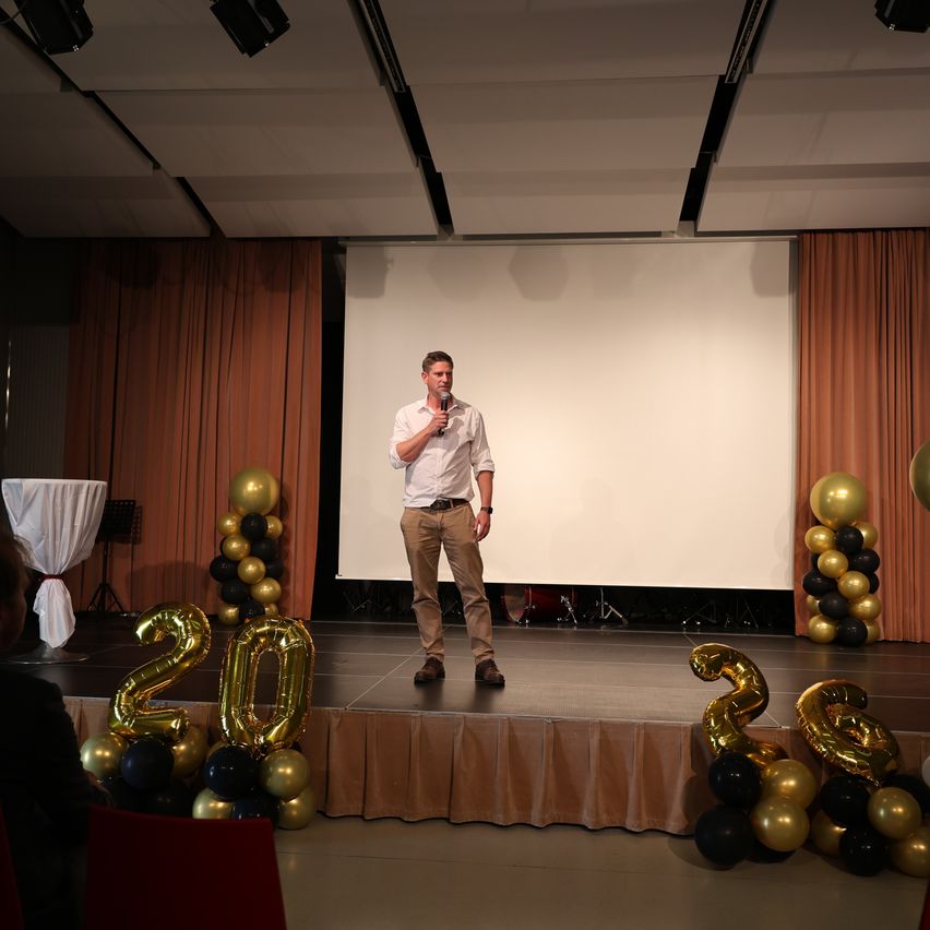 A man stands on a stage with a microphone, likely giving a speech. Behind him is a large white screen, and the stage is decorated with balloons and the number '20'.