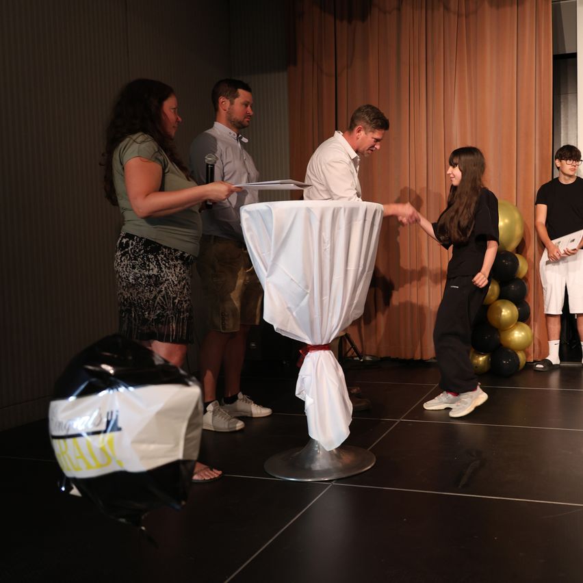 A young woman shakes hands with a man on stage, while a woman holds a microphone and a man holds a paper. Behind them, a table with a white cloth and balloons is visible.