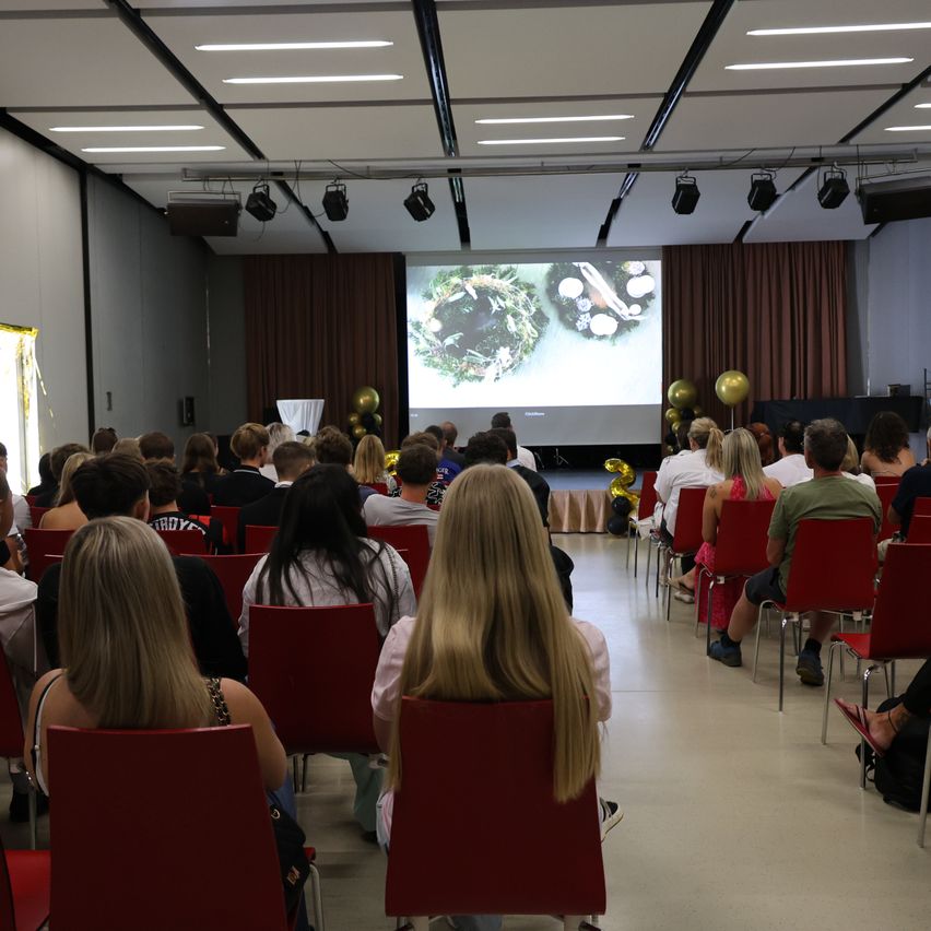 A room filled with people seated in red chairs, watching a presentation on a large screen. The room has a modern design with white walls and ceiling lights.