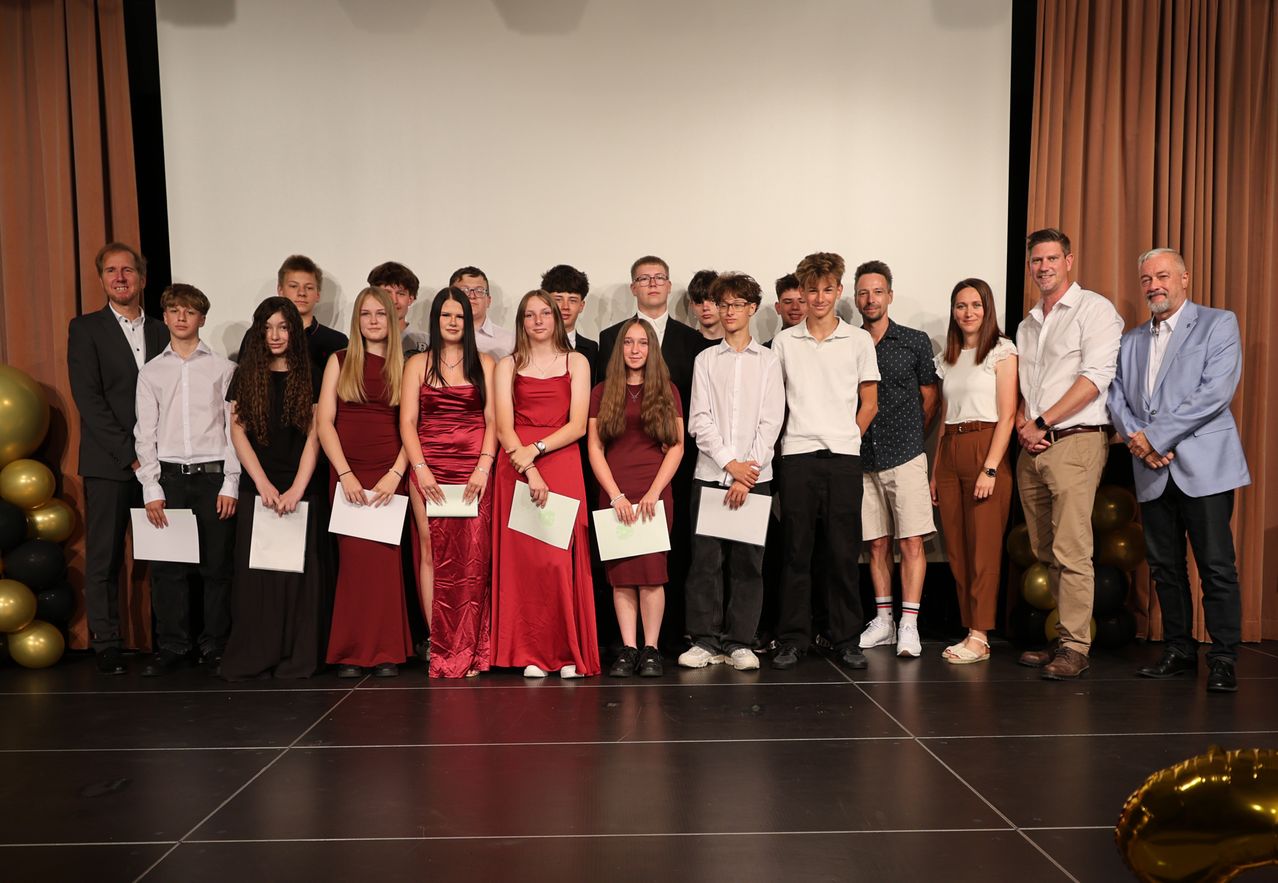 A group of young people, some in red dresses and others in casual attire, stand on a stage holding certificates. They are smiling and appear to be celebrating an achievement.