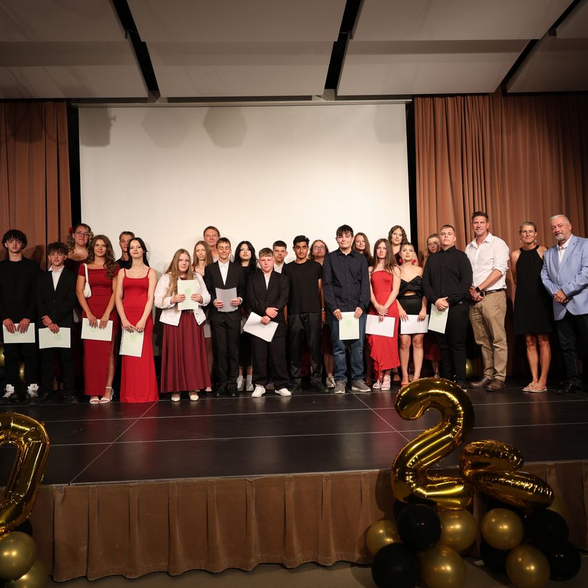 A group of people, likely students, stand on a stage holding certificates. They are surrounded by balloons and a backdrop with a number 25.