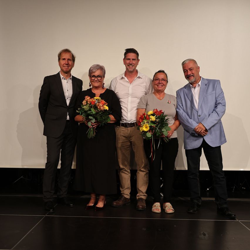 Five individuals stand on a stage, smiling for a photo. Two women hold flower bouquets, one in yellow and the other in a mix of colors. The men wear suits and casual attire.