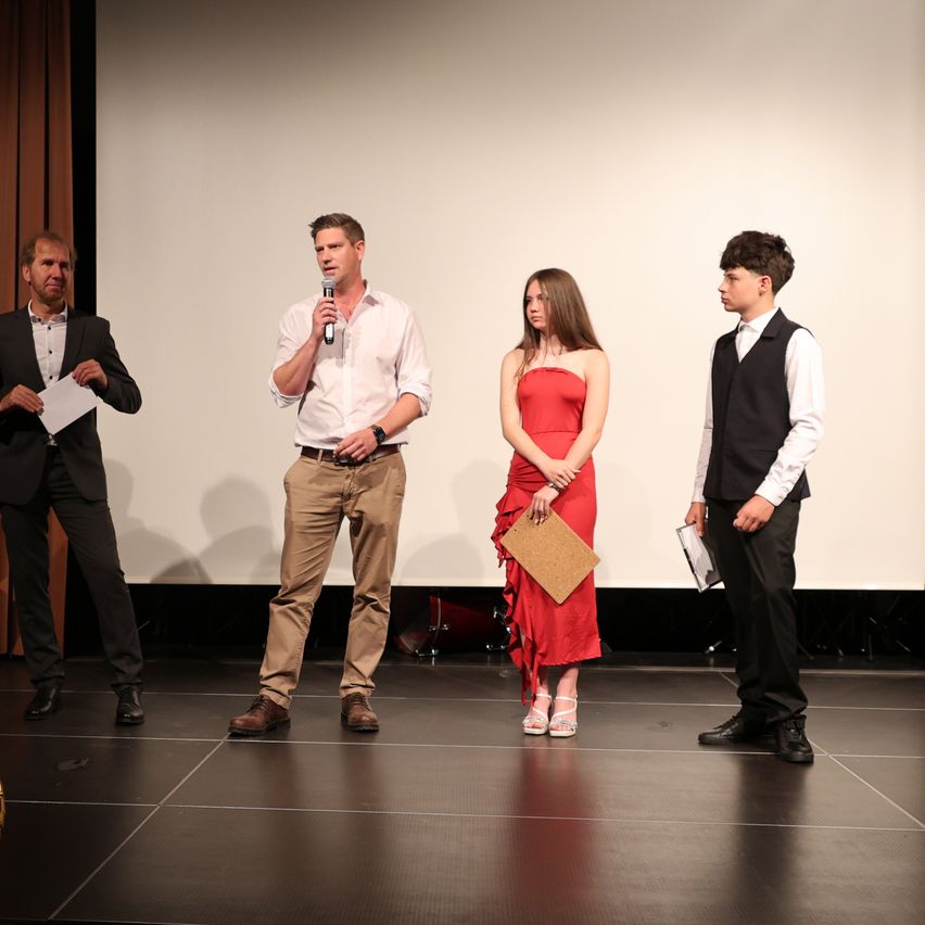 Four individuals stand on a stage, one holding a microphone, another holding a piece of paper, and two others dressed in formal attire. The woman in a red dress holds a folder.